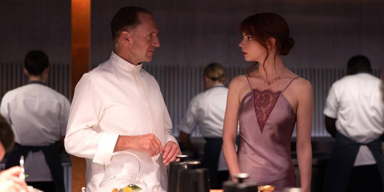 Zu sehen ist ein Mann in einem weißen Kochkittel, der einer Frau in einem eleganten Abendkleid gegenüber steht.